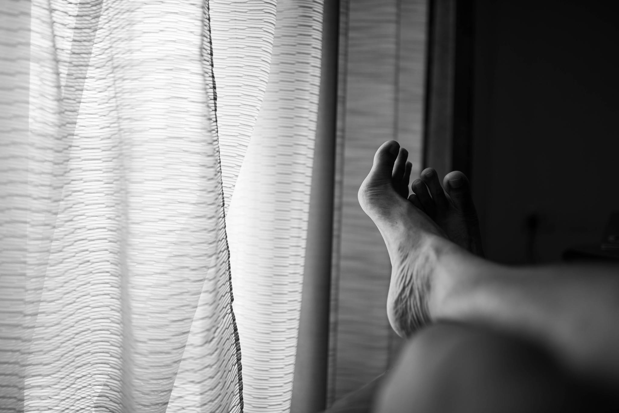 Black and white photo of bare feet in a relaxed pose by a window with curtains.