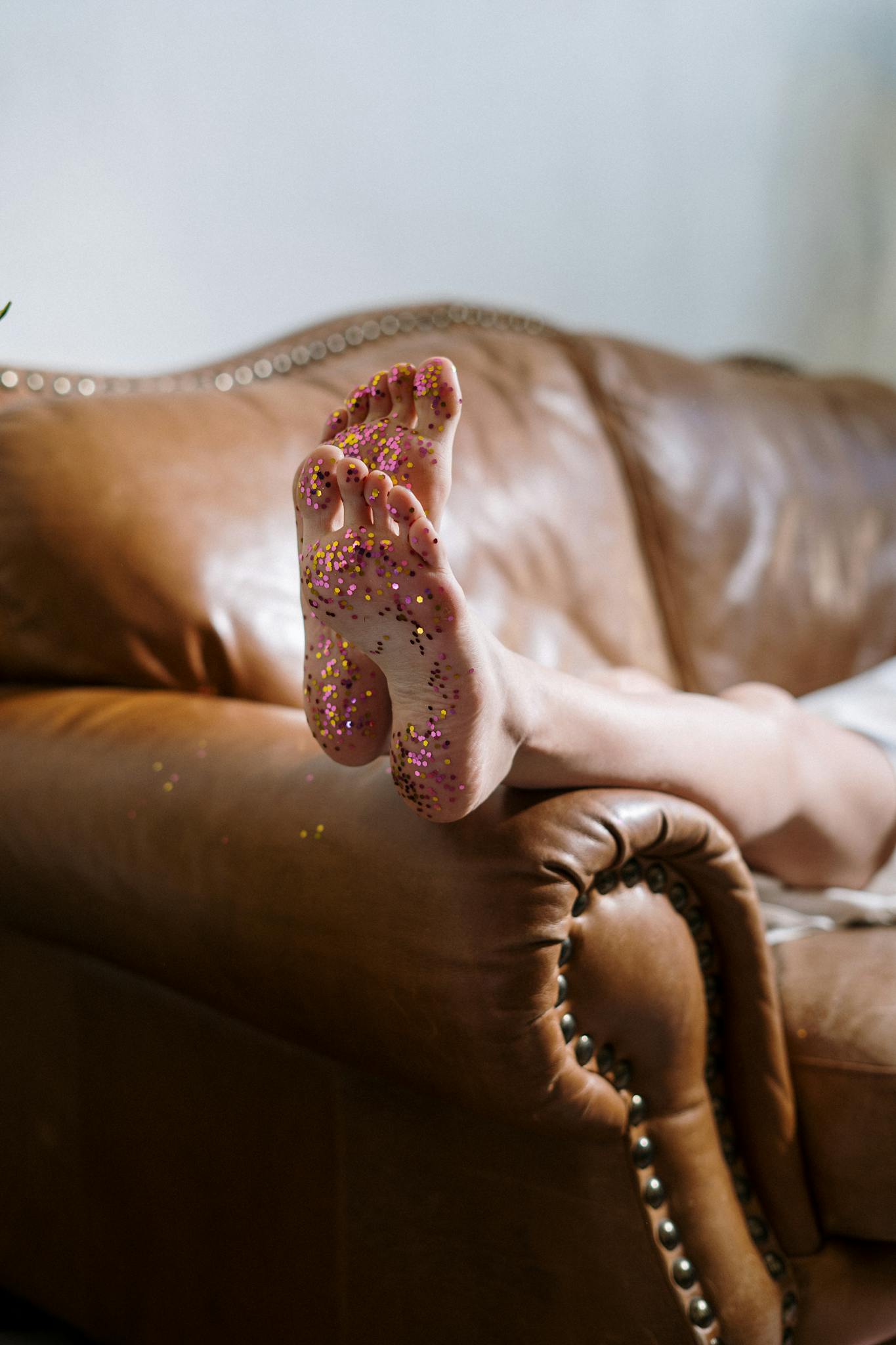 Close-up of feet with glitter, resting on a leather sofa in a stylish setting.